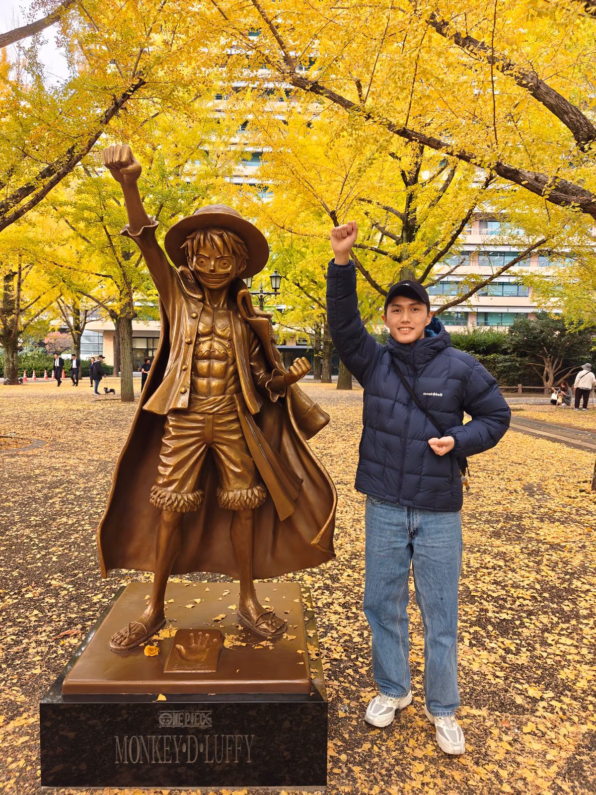 Jun Qi posing beside a Monkey D. Luffy statue under autumn trees